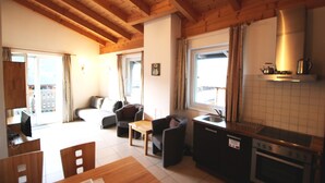 A cozy room with a wooden ceiling, a dining area with a table and chairs, a kitchen with a stove and sink, and a balcony with curtains.