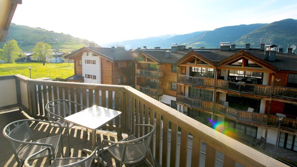 A balcony with a table and chairs, overlooking a mountainous landscape and a row of buildings.