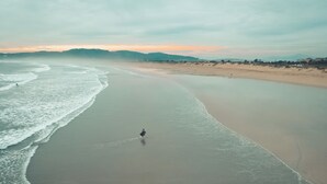 Luchtbeeld van een strand met een persoon die langs de waterlijn loopt, een rustige zee en heuvels in de verte.
