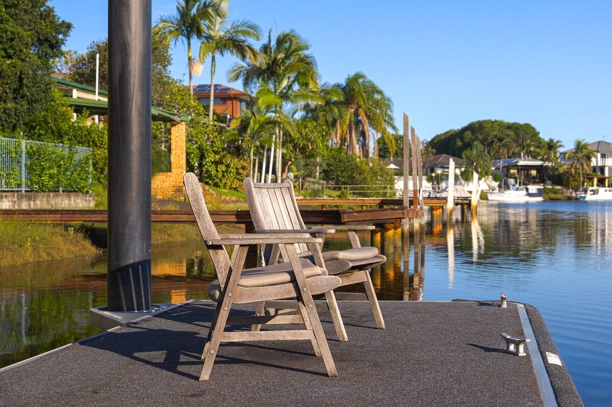 A waterfront area with wooden chairs on a dock, a calm water body, and a backdrop of palm trees and houses.