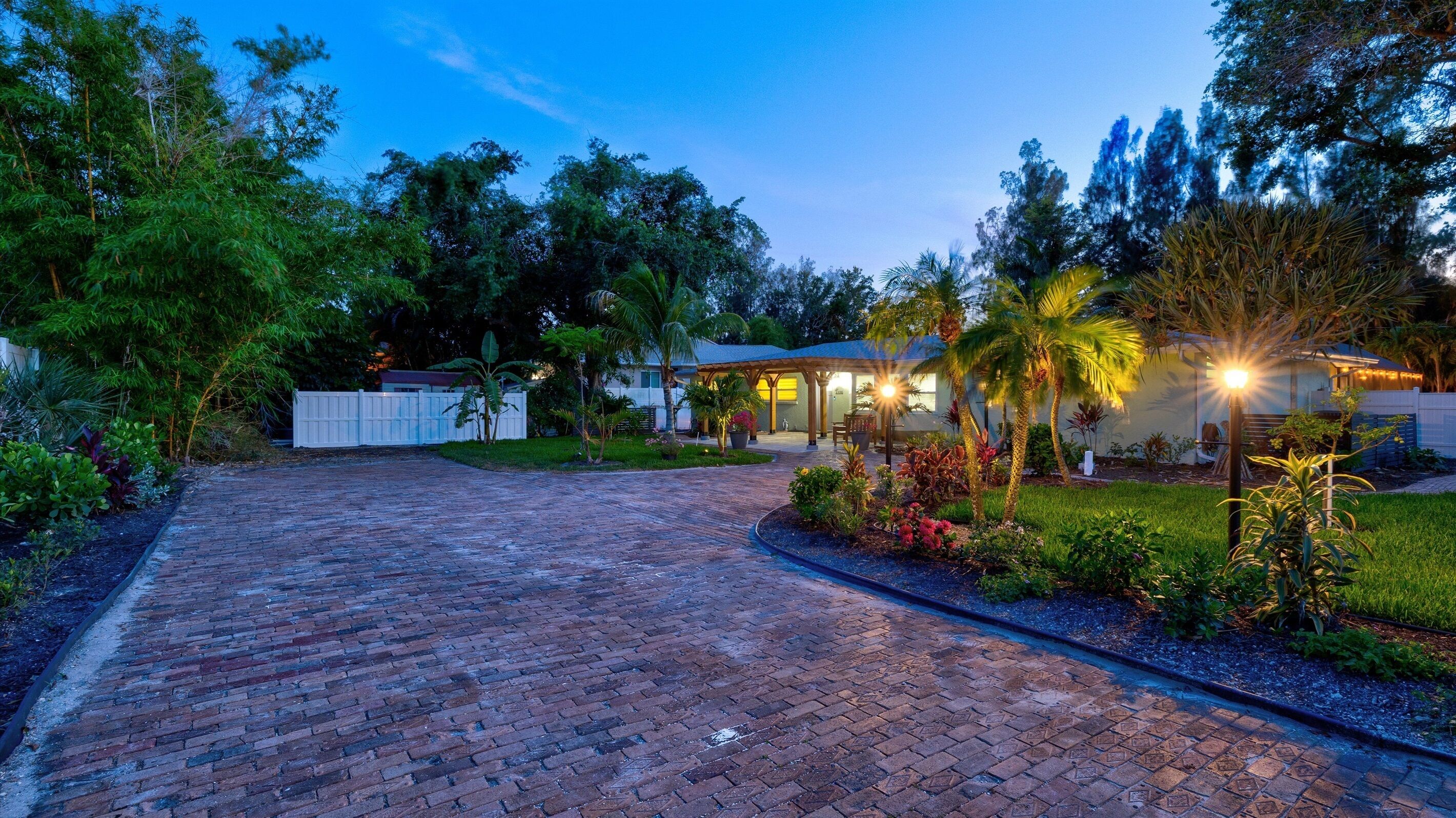 A paved driveway leading to a house with a covered patio area, surrounded by lush greenery and palm trees.