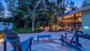 An outdoor patio with Adirondack chairs, a patterned rug, and a pathway leading to a building with glass doors and string lights. - Luxury Getaway | Saltwater Pool, Fire Pit & Games! (Bradenton)