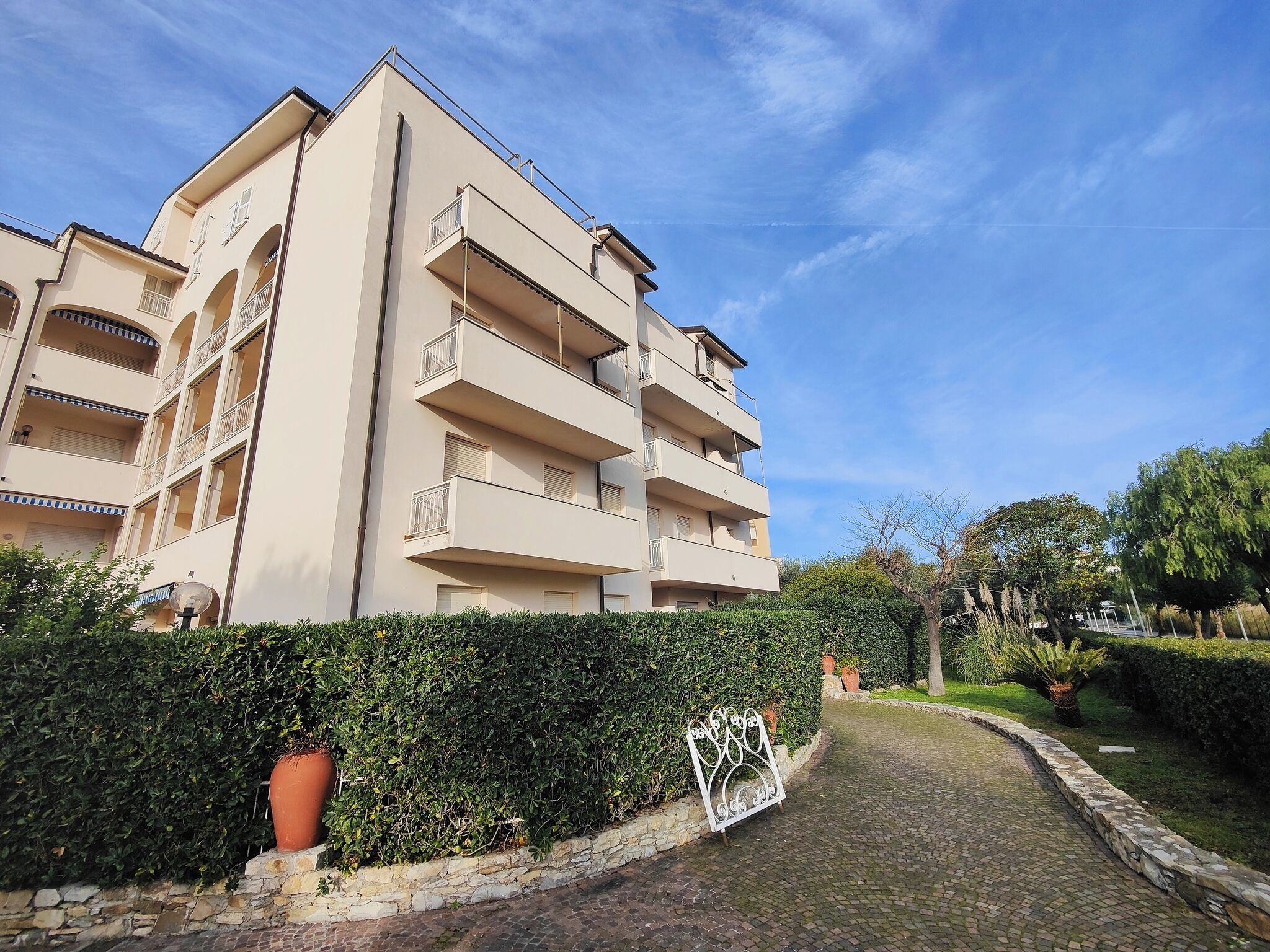 A multi-story residential building with balconies, a paved pathway, and a hedge.