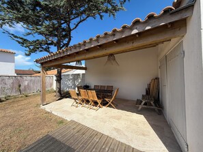 Alpendre coberto com mesa e cadeiras, luminária pendente e um espaço de armazenamento com objetos de madeira.