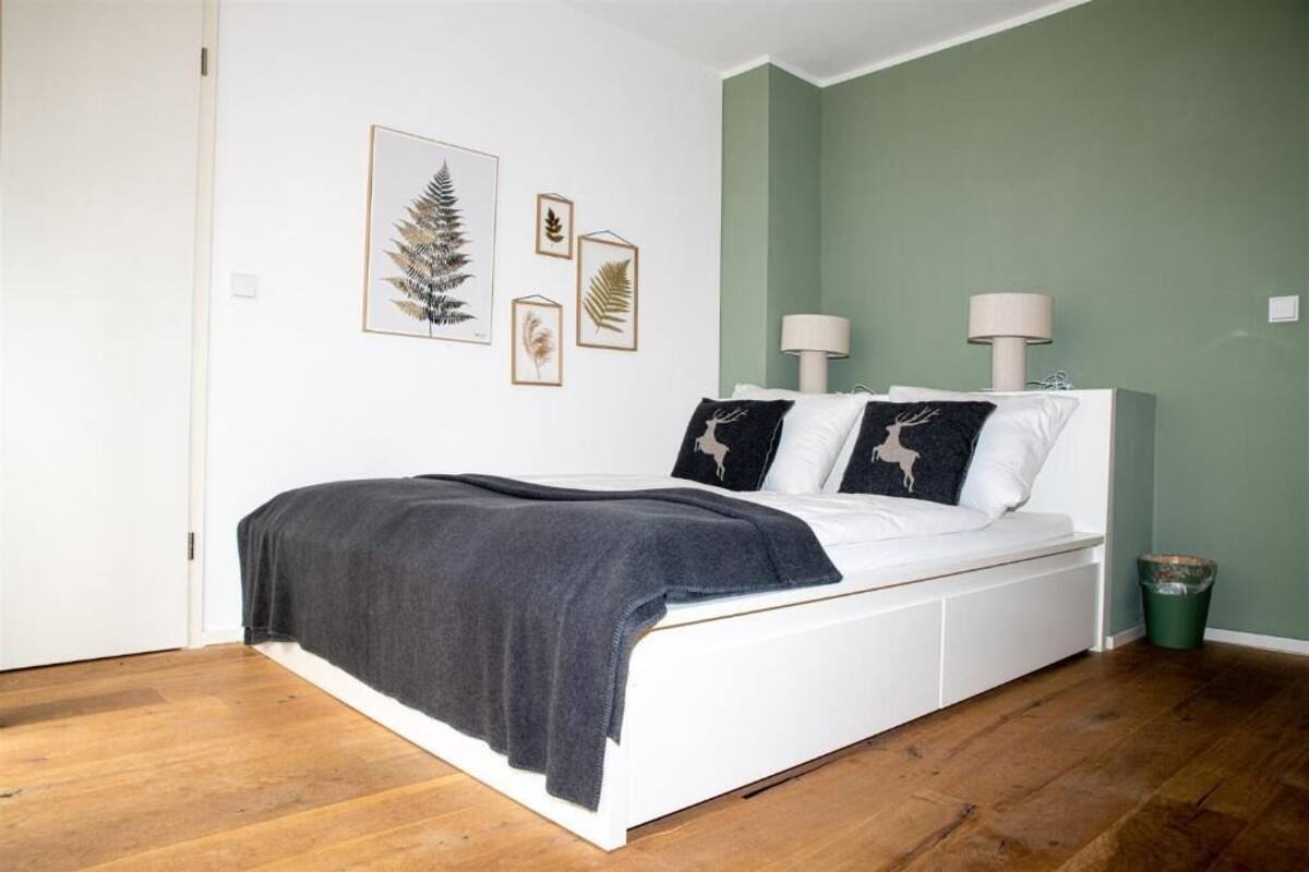 A bedroom with a white bed, a dark grey blanket, and three framed botanical prints on the wall.
