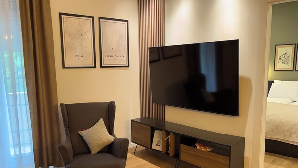 Uma sala de estar moderna com uma televisão de tela plana, uma poltrona escura e um pequeno banquinho. Há dois mapas emoldurados na parede, uma janela com cortinas e uma cama no quarto vizinho.