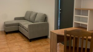 Una sala de estar con un sofá seccional gris, una mesa de comedor de madera y una estantería blanca.