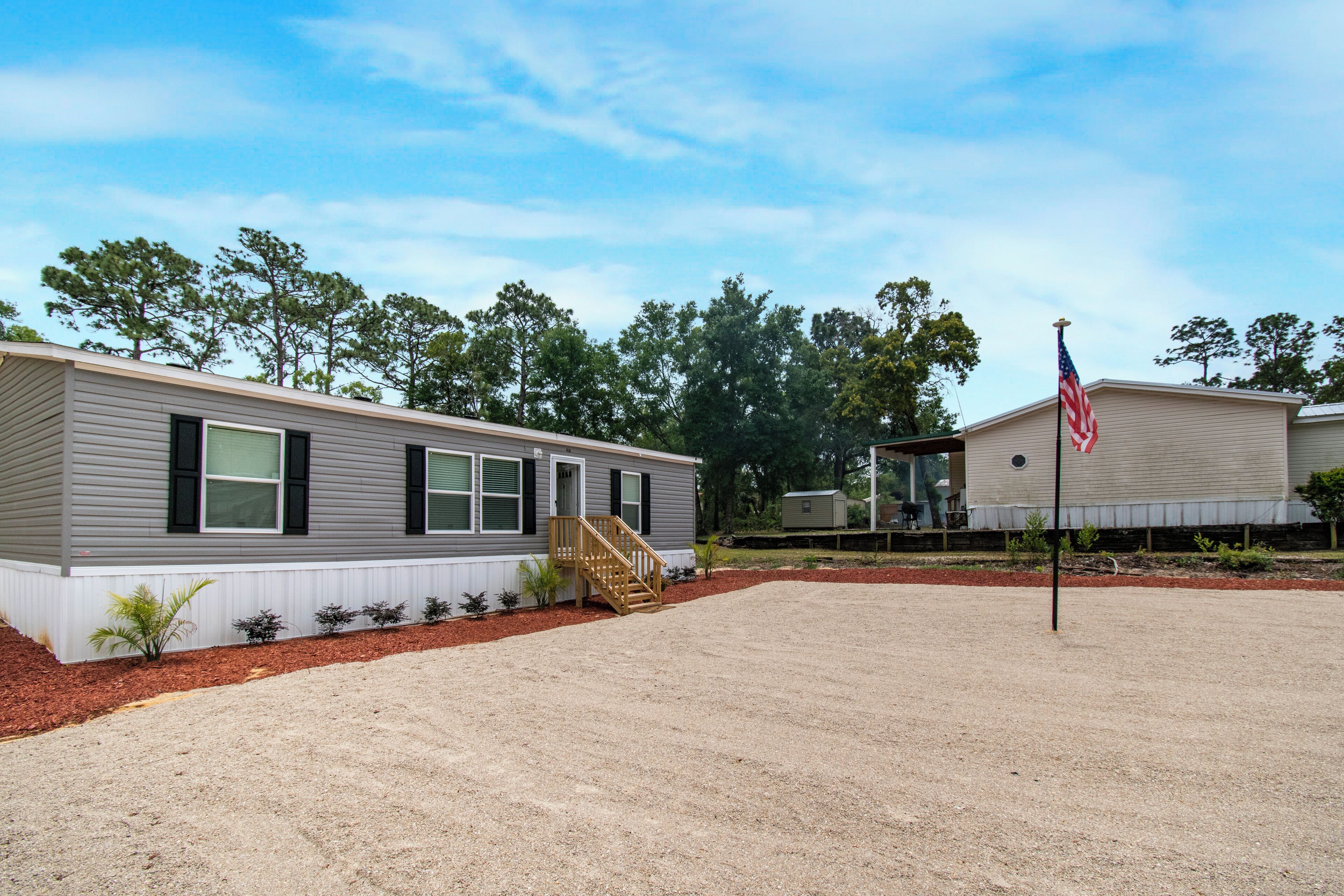 A mobile home with a wooden staircase leading to the entrance, a flagpole with an American flag, and a clear sky.