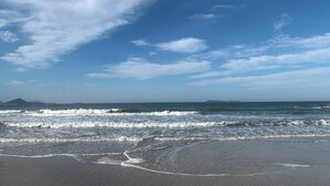 Uma praia com ondas, céu claro e montanhas ao longe.