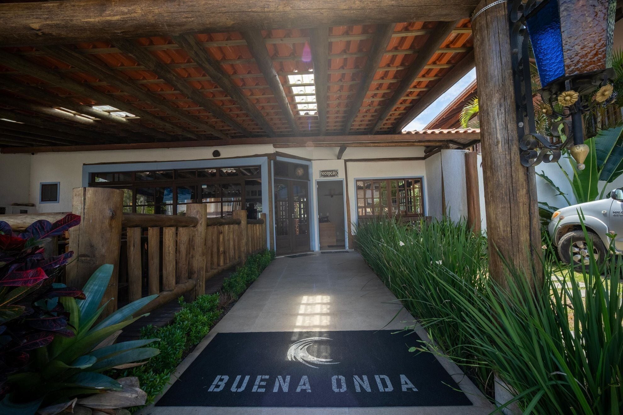 A reception area with a wooden bench, a mat with 'BUENA ONDA' text, and a car parked outside.