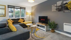 Un salon moderne avec un canapé gris, des coussins jaunes, un téléviseur posé sur un meuble blanc et une grande fenêtre donnant sur des immeubles.