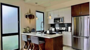 A modern kitchen with a central island, bar stools, stainless steel appliances, and pendant lighting. - Mision Viejo Sur 1BR Apartment B103 (Playas de Rosarito)