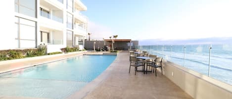 A rooftop pool with a view of the ocean, surrounded by lounge chairs and a building with multiple floors.