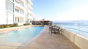 A rooftop pool with a view of the ocean, surrounded by lounge chairs and a building with multiple floors.