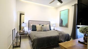 A modern hotel room with a large bed, bedside tables with lamps, a ceiling fan, a large window with curtains, and a painting on the wall.