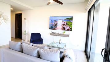 Una sala de estar moderna con un sofá gris, un sillón azul, una mesa de centro de vidrio y un televisor de pantalla plana montado en la pared que muestra un paisaje pintoresco.