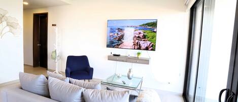 A modern living room with a grey sofa, a blue armchair, a glass coffee table, and a flat-screen TV mounted on the wall displaying a scenic landscape.