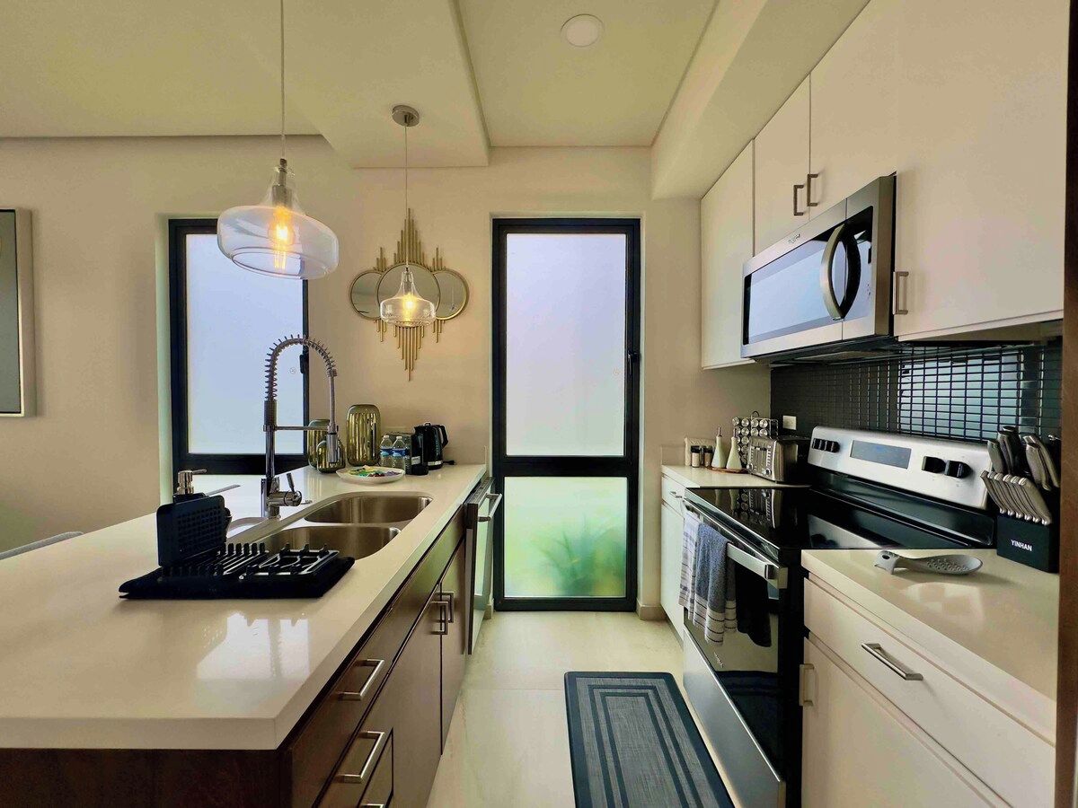 A modern kitchen with a central island, stainless steel appliances, and a view through a large window.
