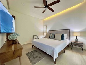 A modern bedroom with a large bed, a ceiling fan, a TV mounted on the wall, and a bedside table with a lamp.
