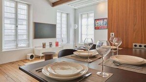 Un coin repas avec une table en bois dressée pour quatre personnes, un téléviseur à écran plat fixé au mur et de grandes fenêtres équipées de stores.