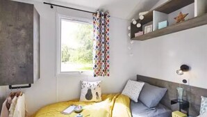 A bedroom with a bed, a window with curtains, a shelf with decorative items, and a bedside table with a lamp.