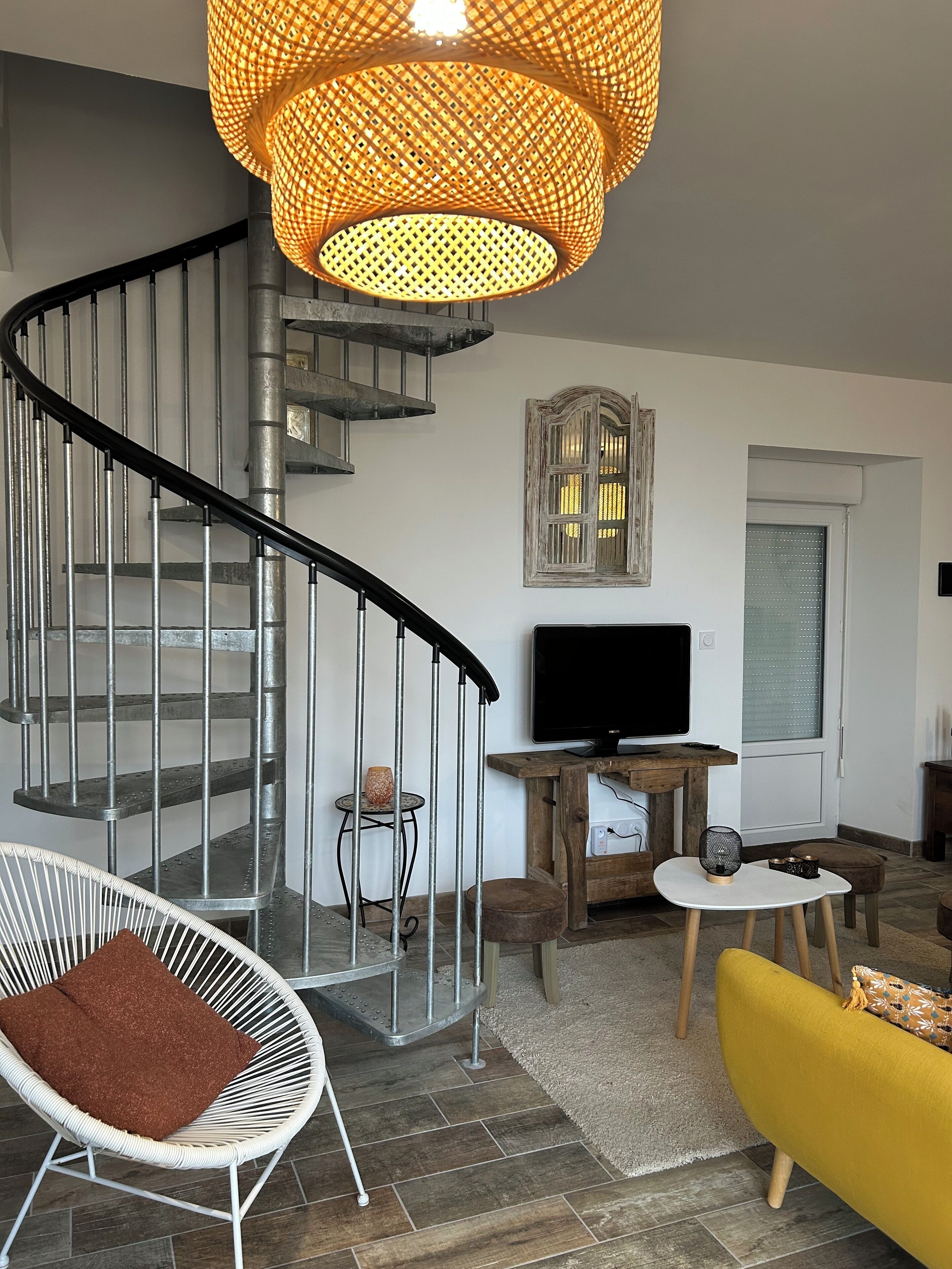 Una sala de estar moderna con una escalera espiral, un televisor de pantalla plana, una silla blanca, un sofá amarillo y una lámpara colgante grande de fibras naturales.