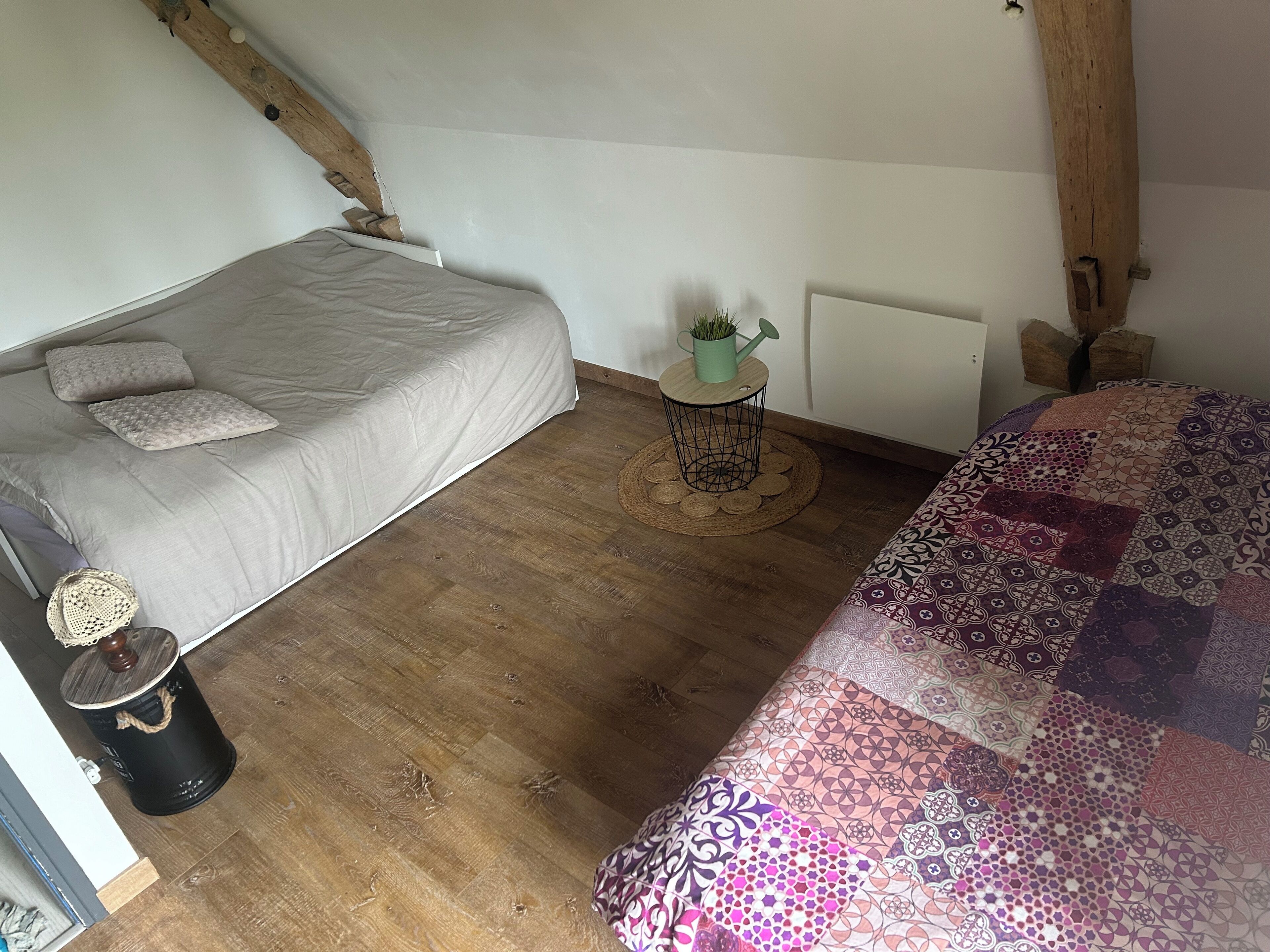 Ett sovrum med trägolv, en säng med ett beige överkast, ett litet runt bord med en växt och ett element.