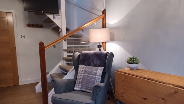 Una sala de estar con un sillón azul, un banco de madera, una escalera y un televisor montado en la pared.