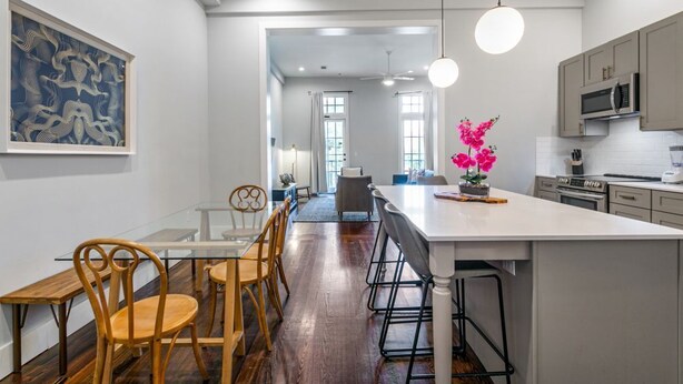 A modern kitchen with a large island, wooden chairs, and a dining table. There is a painting on the wall and a vase with flowers on the island. - Luxurious 5bd with private balcony/ AMAZING LOCATION! (New Orleans)