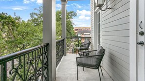 A covered porch with two chairs and a table, a black wrought iron railing, and a view of trees and buildings in the background. - Luxurious 5bd with private balcony/ AMAZING LOCATION! (New Orleans)