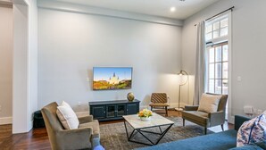 A spacious living room with a large window, a flat-screen TV mounted on the wall, a dark-colored entertainment unit, and a comfortable seating area with a sofa and two armchairs. - Luxurious 5bd with private balcony/ AMAZING LOCATION! (New Orleans)
