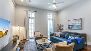 A spacious living room with a large window, a ceiling fan, a blue sofa, and a coffee table with flowers. - Luxurious 5bd with private balcony/ AMAZING LOCATION! (New Orleans)