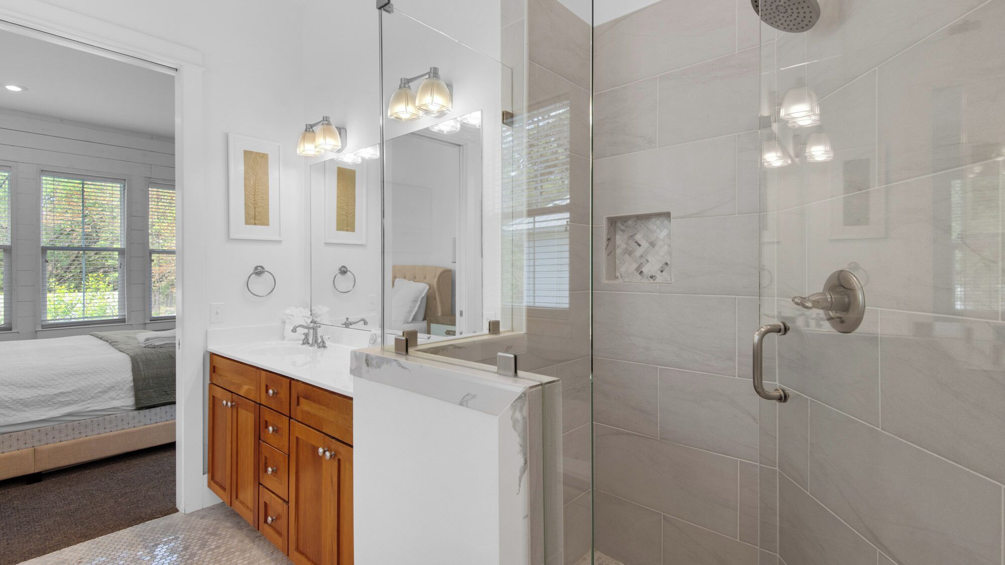 A modern bathroom with a glass shower enclosure, a vanity with a mirror, and a bed in the adjacent room.