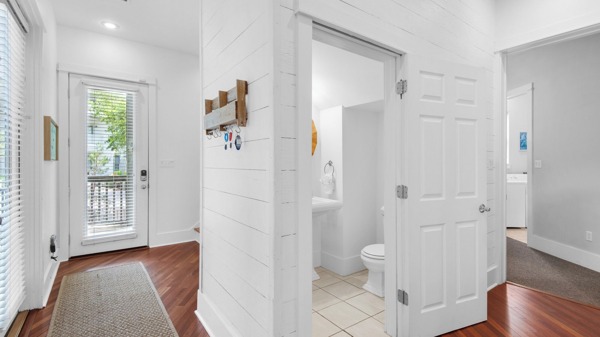 Een gang met een houten vloer, witte muren en een deur die leidt naar een badkamer met toilet en wastafel.