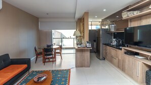 Una sala de estar moderna con sofá, una mesa de centro y un comedor con sillas. Cuenta con una cocina, un refrigerador, un microondas y un televisor de pantalla plana grande.