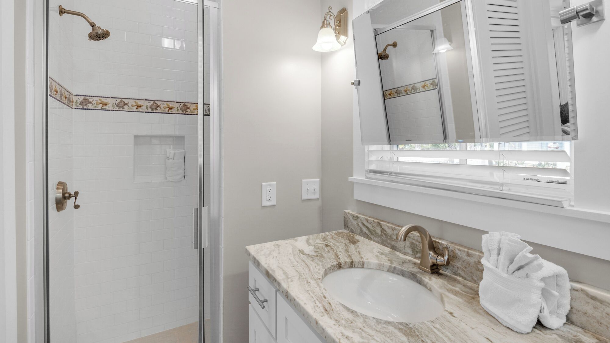 Une salle de bain dotée d’une porte de douche en verre, d’un lavabo avec un plan de toilette en marbre et d’un miroir au-dessus du lavabo.