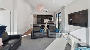 Una sala moderna con sofá, dos sillones, televisión y una cocina al fondo.