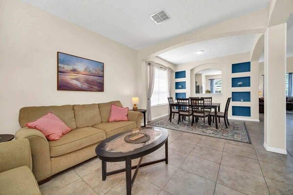 A living room with a beige sofa, a coffee table, and a painting on the wall. There is a dining area with a table and chairs, and a kitchen area with blue cabinets. - Remodeled 7 Bedroom Retreat Near Disney World (Kissimmee)