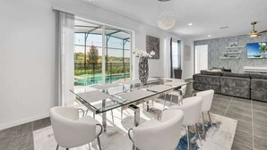 A modern dining area with a glass table and white chairs, a gray sofa, and a large window with a view of greenery. - *NEW* Super Amazing Vacation Getaway (Kissimmee)