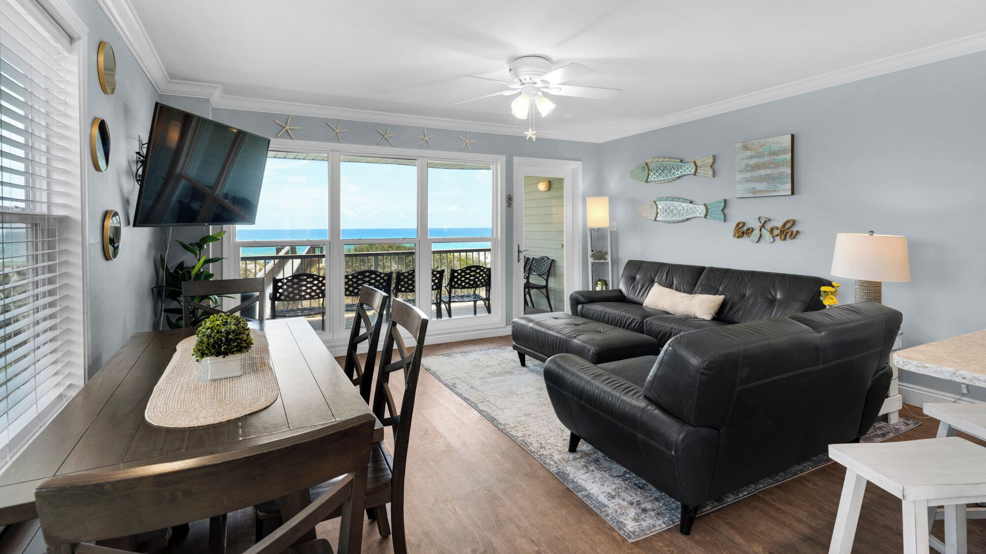 A living room with a black leather sofa, a dining table with chairs, a TV mounted on the wall, and a view of the ocean through large sliding glass doors.