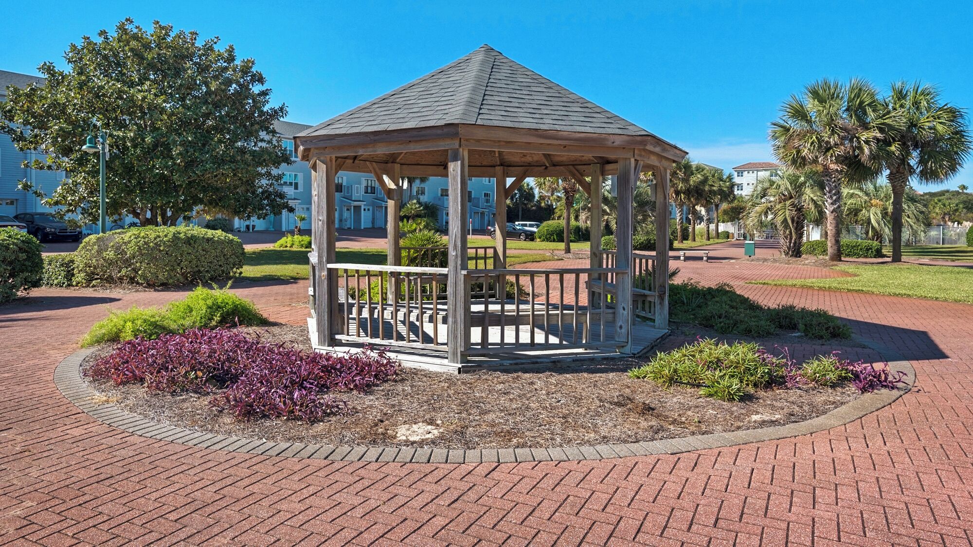 A gazebo with a wooden structure and a roof, surrounded by a brick pathway and landscaped areas with shrubs and a palm tree.