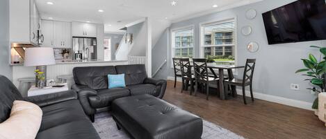 A modern living room with a black leather sectional sofa, a dining area with a table and chairs, and a kitchen in the background.