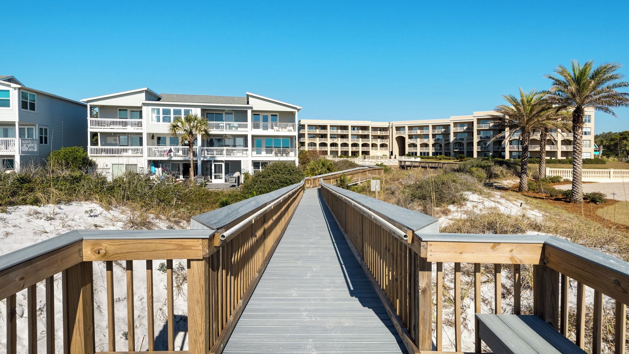 A wooden boardwalk leading to a beachfront hotel with multiple balconies and a sandy beach.