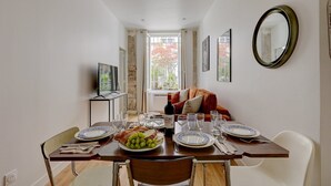 A dining area with a long table set for a meal, a television on a stand, and a window with a view of greenery. - 397 Suite Glamour - Superb Studio in Paris (Paris 10 Entrepôt)