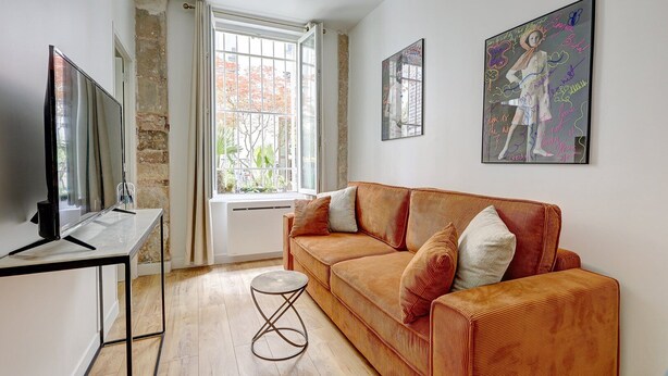 A living room with a sofa, a TV, a window, and artwork on the walls. - 397 Suite Glamour - Superb Studio in Paris (Paris 10 Entrepôt)