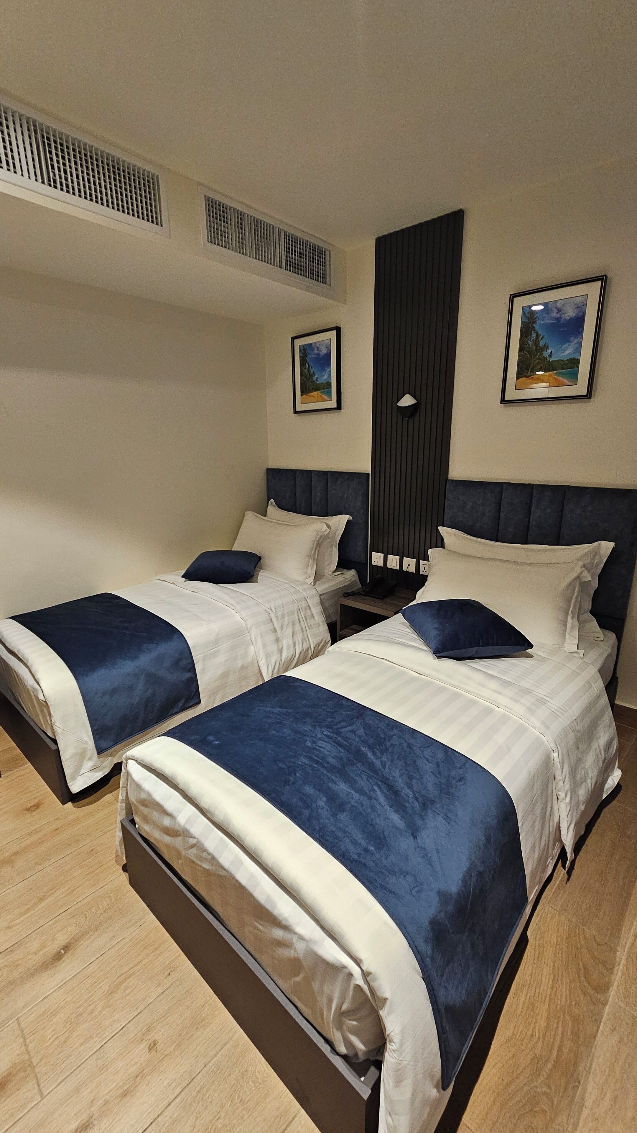 A hotel room with two beds, a dark headboard, and a wall-mounted air vent.