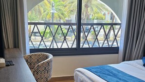 Une chambre d’hôtel avec un lit, un bureau, une chaise et une grande fenêtre donnant sur des palmiers et une clôture.