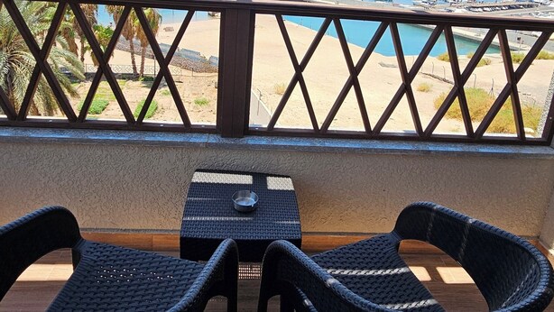 Un balcon avec des fauteuils et une table en osier, donnant sur une plage et une marina où sont amarrés des bateaux.