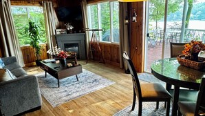 Een gezellige woonkamer met houten vloeren en een open haard, een eethoek met een glazen tafel, en grote ramen met uitzicht op de omgeving.
