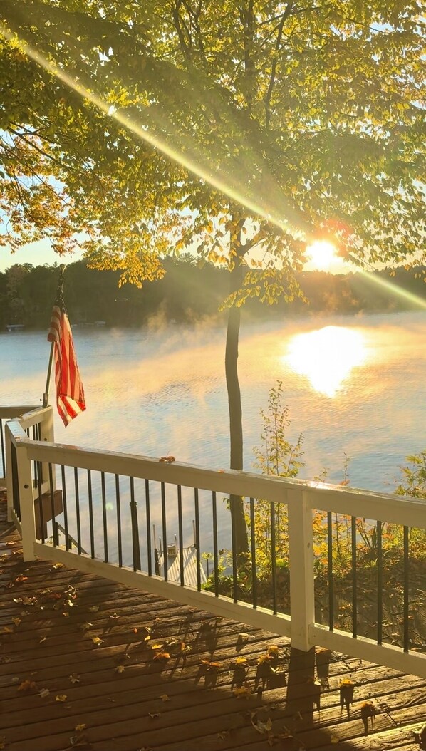 A wooden deck with a railing overlooking a calm lake, with sunlight filtering through trees.